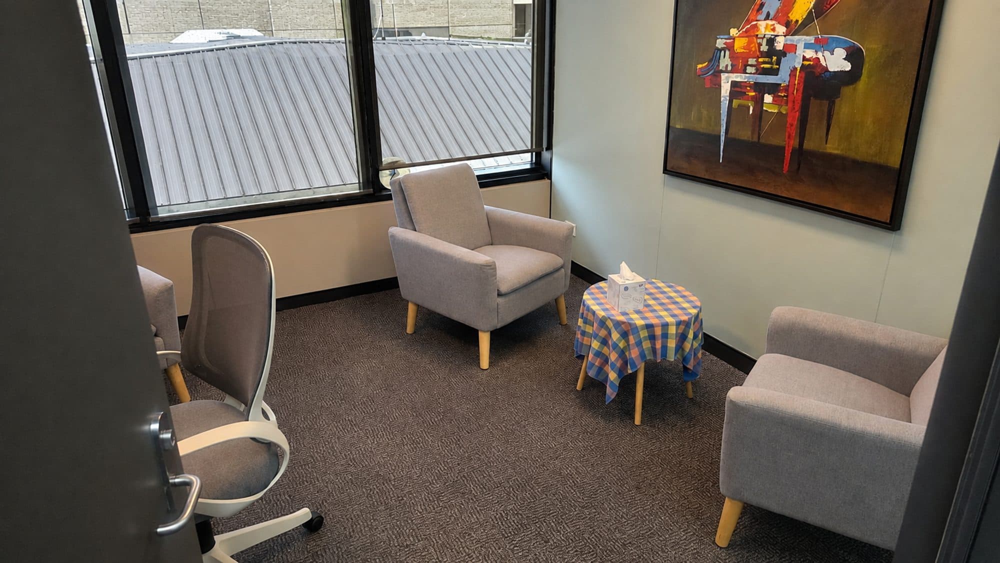 The counselling room at The Listening Room in Curtin: two soft armchairs facing each other, a small table with a box of tissues, a window, and a framed painting on the wall.